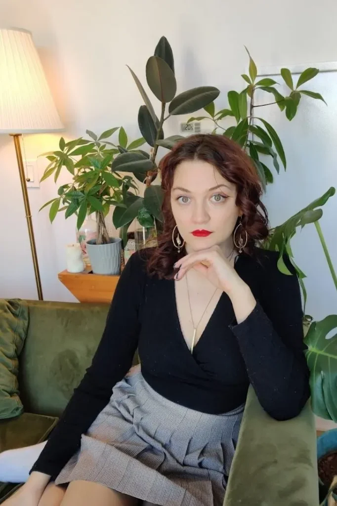 Megan, a white femme with brown hair, red lipstick stares at the camera. She is sitting on a green couch, behind her are large plants